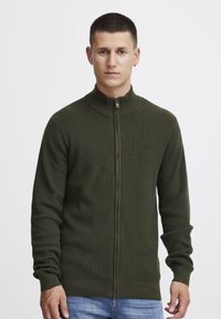 Olivengrønn ribbet zip-up genser med høy krage, tettsittende passform og lange ermer. Har en sølvfarget glidelås og teksturert stoff.
