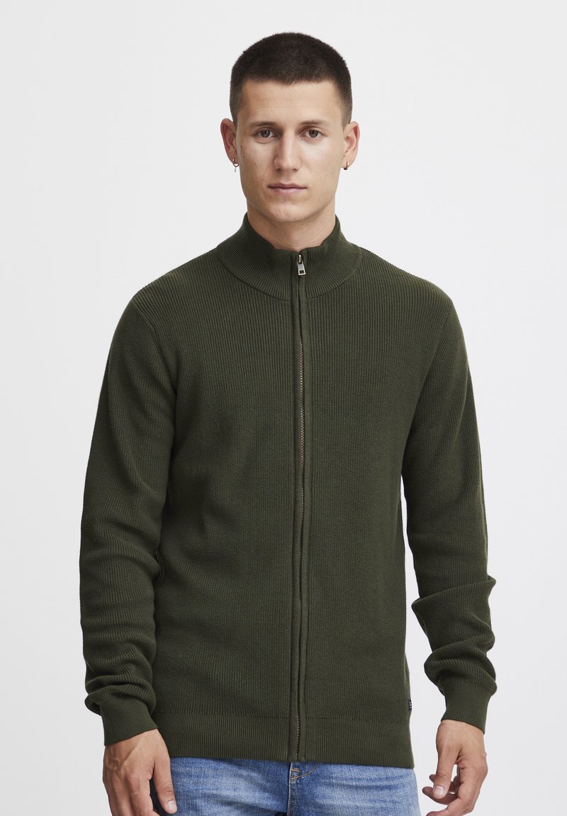 Pull en maille côtelée vert olive à zip avec col montant, coupe ajustée et manches longues. Présente une fermeture éclair argentée et un tissu texturé.