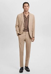 Man stands wearing a beige suit with a brown polo shirt and black dress shoes against a plain light background.