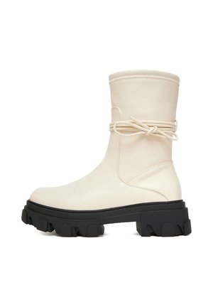 Kremowy but do połowy łydki z grubą czarną podeszwą typu lug oraz skórzanym sznurowaniem owiniętym wokół buta i zawiązanym na kokardę z boku.