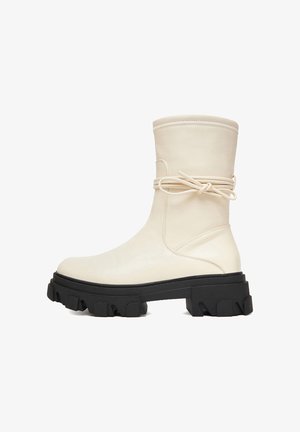 Bota color crema hasta media pantorrilla con suela gruesa negra de perfil dentado y cordones de cuero que se envuelven alrededor y se atan en un lazo en el lateral.