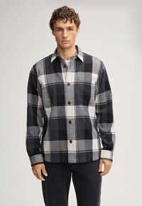 Jonge man in een zwart, wit en grijs groot geruit overhemd met donkere broek, staande tegen een effen lichte achtergrond.