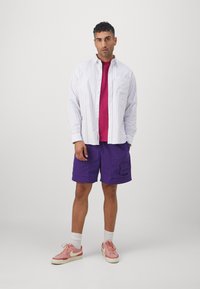 Stribet hvid skjorte over en magentafarvet t-shirt, kombineret med lilla shorts og lyserøde sneakers. Funktioner inkluderer sidelommer og en afslappet pasform.