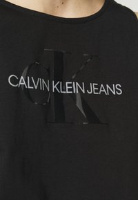 Schwarzes Baumwoll-Tanktop mit einem glänzenden "OK"-Logo und dem weißen Text "CALVIN KLEIN JEANS". Glatte Textur, lässiges Design.