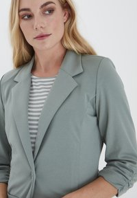 Ljusgrön blazer med en figursydd design, som har en notch-krage och en enkel knappstängning, lager på lager över en grå- och vitrandig topp.