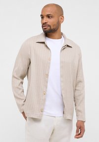 Giacca beige a righe con colletto, chiusura con bottoni e maniche lunghe, indossata sopra una camicia bianca e pantaloni di colore chiaro.