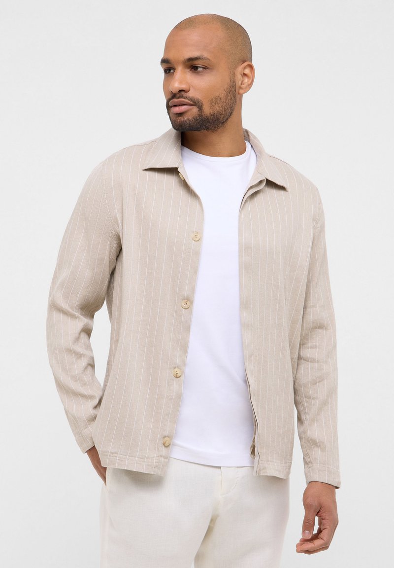 Giacca beige a righe con colletto, chiusura con bottoni e maniche lunghe, indossata sopra una camicia bianca e pantaloni di colore chiaro.