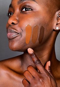 Tre färgprover av foundationnyanser på mörk hud, som varierar från ljus till mörk brunt. Modellen visar en finger som pekar på färgproverna.