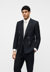 Άνδρας που φοράει σφιχτά εφαρμοστό μαύρο κοστούμι διπλής γραμμής με λευκό πουκάμισο και ανοιχτό μπεζ γραβάτα σε απλό λευκό φόντο.