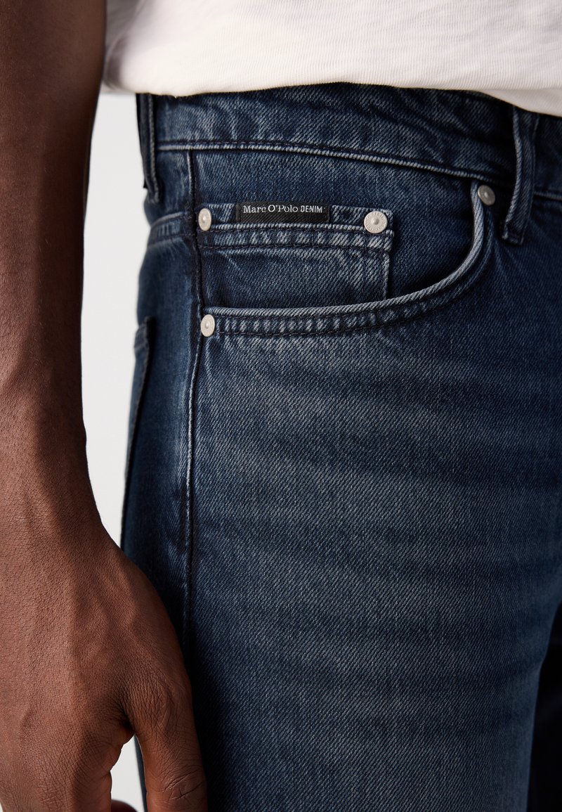 Granatowe dżinsy mają dopasowany krój, widoczne szwy, metkę projektanta na tylnej kieszeni oraz metalowe akcenty w postaci guzików.