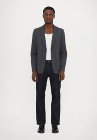 Grauer Blazer mit Reverskragen, Vorderaschen und taillierter Passform, kombiniert mit einem weißen Top und dunklen Jeans. Schwarze Schuhe.