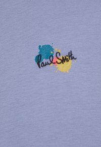 Tissu bleu clair avec texte cursif noir "Paul Smith" sur des motifs éclaboussés de peinture bleue, rouge et jaune brodés sur la surface.