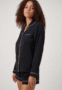 Svart pyjamasset som innehåller en långärmad skjorta med knappar och vit kantning, samt matchande shorts med samma detalj, tillverkat av mjukt tyg.