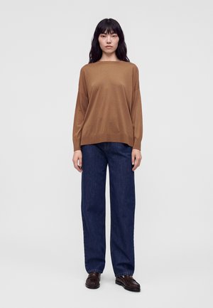 Frau, die steht, trägt einen braunen Langarmpullover, dunkelblaue Hosen mit weitem Bein und dunkelbraune Loafers vor einem schlichten hellen Hintergrund.