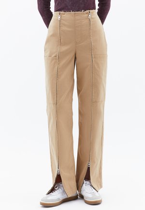 Beige cargobroek met twee zijzakken, zichtbare ritsen aan de zijkanten en een afgewassen taillebanddetail. Gecombineerd met witte sneakers.