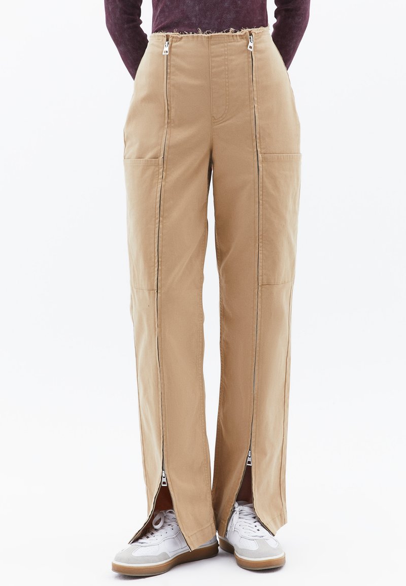 Beige cargobroek met twee zijzakken, zichtbare ritsen aan de zijkanten en een afgewassen taillebanddetail. Gecombineerd met witte sneakers.
