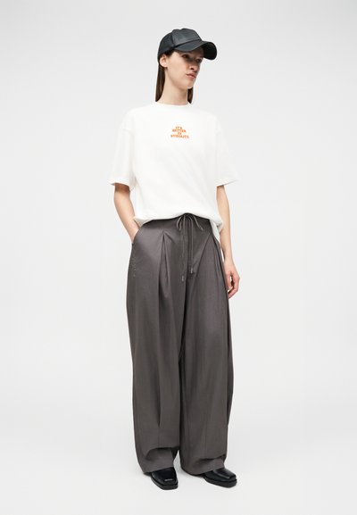 Γυναίκα με λευκό φαρδύ t-shirt με πορτοκαλί κείμενο, χαλαρό γκρι πλισέ παντελόνι, μαύρα παπούτσια και μαύρο δερμάτινο καπέλο του μπέιζμπολ, όρθια με τα χέρια στις τσέπες.