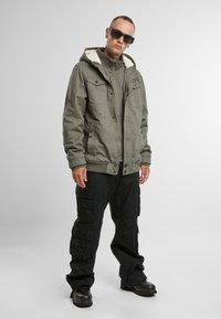 Chaqueta verde oliva con capucha forrada de piel, cierre con botones y cremallera, y múltiples bolsillos, combinada con pantalones cargo negros y botas negras.