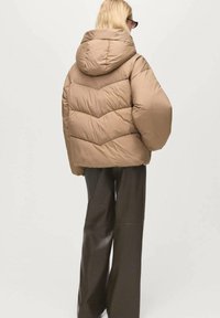 Giacca puffer marrone con cappuccio, caratterizzata da un design a rombi e una vestibilità rilassata. Abbinata a pantaloni scuri a gamba larga.