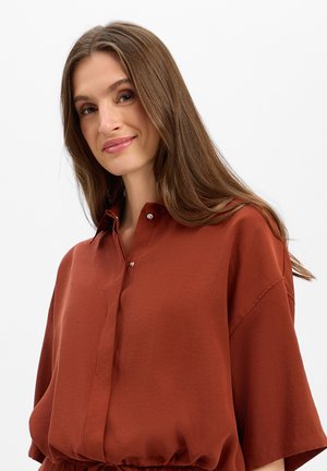 Vrouw met lang bruin haar die een losse roestkleurige blouse met knopen draagt, licht glimlachend tegen een effen lichte achtergrond.