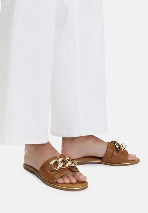 Brązowe skórzane klapki na płaskiej podeszwie z ozdobnym złotym łańcuchem z przodu, z minimalną liczbą pasków, zestawione z białymi spodniami o szerokich nogawkach.