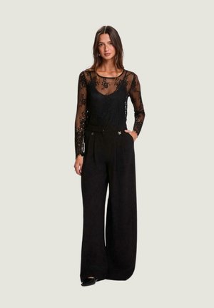 Top de encaje negro de manga larga sobre un camisola negra, combinado con pantalones de pierna ancha y talle alto en negro. Patrón floral sutil en el top.