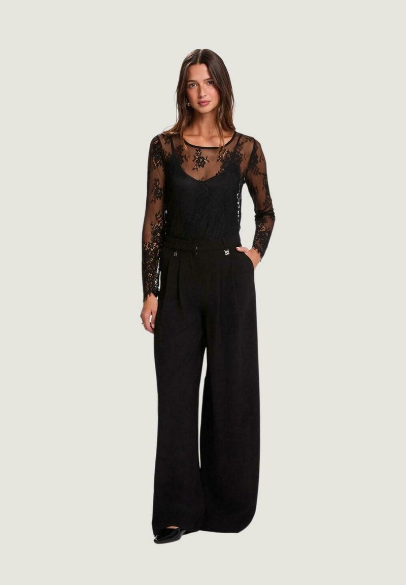 Top de encaje negro de manga larga sobre un camisola negra, combinado con pantalones de pierna ancha y talle alto en negro. Patrón floral sutil en el top.