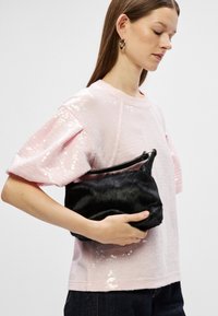 Mustast kunstkarvast kotist kaarjas kuju ja musta tõmblukuga, hoituna heleda roosa sädelevast pluusist, millel on struktuursed varrukad.