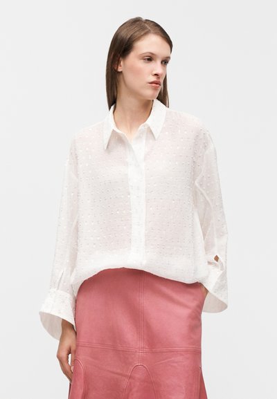 Blouse blanche transparente avec motifs en points texturés, manches longues et col pointu, associée à une jupe en cuir rose.