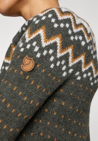 Green knitted sweater featuring a patterned yoke in white and orange, with a small brown leather logo patch on the shoulder.