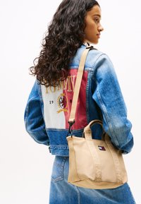 Beige tote bag med to korte håndtag og et langt rem, med tekstureret materiale og præget "TOMMY JEANS"-detaljer. Denimjakke med farverig branding.