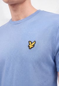 Homme portant un t-shirt bleu clair avec un petit emblème d'oiseau jaune sur la partie gauche de la poitrine.