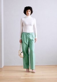White long-sleeve turtleneck, green loose pants, and beige handbag with chain strap. Strappy sandals complete the outfit against a neutral backdrop.