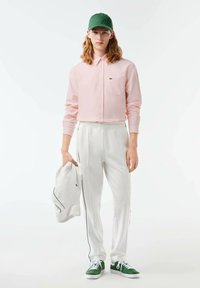 Camicia button-up rosa chiaro, pantaloni tuta bianchi con strisce laterali nere, cappellino verde, mentre tiene una giacca bianca, e sneakers verdi.