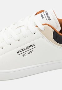 White leather sneaker with white laces, brown and black accents, black logo "JACK & JONES EST. 1990" on the side, and smooth texture.