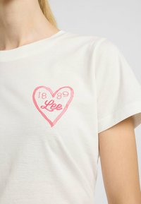 Hvid bomulds T-shirt med et pink hjerte design og teksten "1889" og "Lee" indeni. Korte ærmer og en klassisk rund hals.