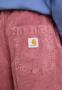 Fakó rózsaszín cordúrnadrág, hátoldalán fehér címkével és narancssárga logóval rendelkező zsebbel. Strukturált anyag, függőleges bordákkal.