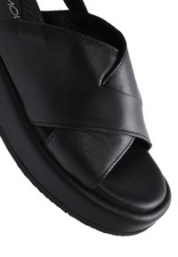 Sandalo in pelle nera con suola a piattaforma spessa. Presenta fascette incrociate, punta arrotondata e una texture liscia. Dettagli di cucitura robusta visibili.