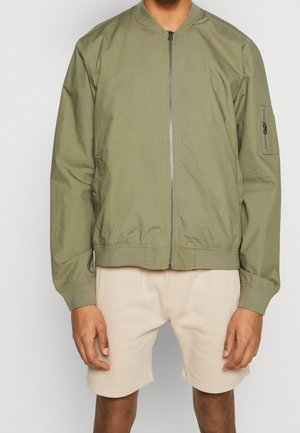Homme portant une veste bomber kaki zippée avec un short beige, debout, les bras détendus le long du corps, devant un fond uni.