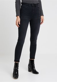 Černé skinny džíny s vysokým pasem, které mají přední kapsy, zapínání na knoflík a prošité detaily. Kombinovány s lesklými černými lodičkami na kotníku.