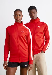 Røde zip-up jakker med høje kraver, hvide side-striber, ribbede manchetter og logo detalje. Den ene model har shorts på, mens den anden har hænderne i lommerne.