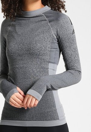 Haut de sport gris à manches longues avec un motif texturé, col haut et trous pour les pouces. Présente un logo discret sur l'épaule.
