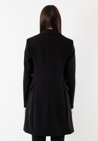 Piovera lunga nera con un design su misura, colletto a revers, tasche laterali e una texture liscia, vista da dietro su uno sfondo bianco.