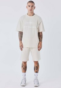 Chemise crème à manches courtes avec un grand logo blanc, assortie à un short. Tissu texturé, coupe décontractée, tatouages accentués et baskets.