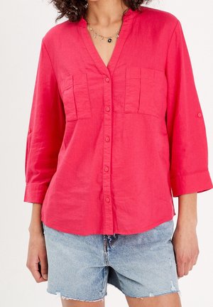 Femme portant une chemise rose vif boutonnée avec deux poches poitrine et un short en jean bleu clair aux ourlets effilochés.