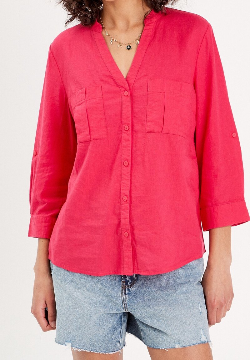 Femme portant une chemise rose vif boutonnée avec deux poches poitrine et un short en jean bleu clair aux ourlets effilochés.