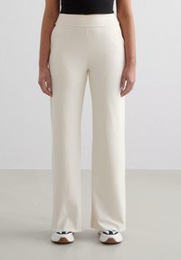 Brede broek in zachte crème stof met een gladde textuur, hoge taille en geen zichtbare sluitingen, gecombineerd met witte sportschoenen.