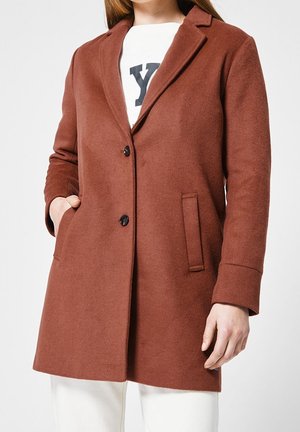 Femme portant un manteau en laine couleur rouille avec deux boutons noirs, col cranté et poches latérales, par-dessus une chemise blanche avec des lettres noires.