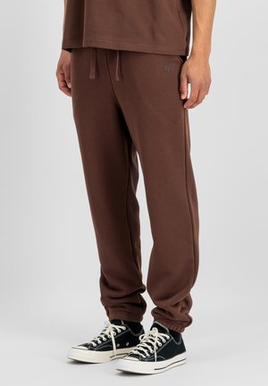 Mann trägt braune Jogginghose mit elastischen Bündchen und schwarze flache Canvas-Sneaker, steht vor einem schlichten grauen Hintergrund.