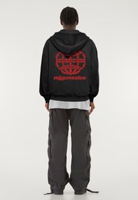 Fekete kapucnis pulóver piros grafikával a hátán, melyen a "mjgonzales" felirat található, laza fitnesz szürke cargo nadrággal párosítva.